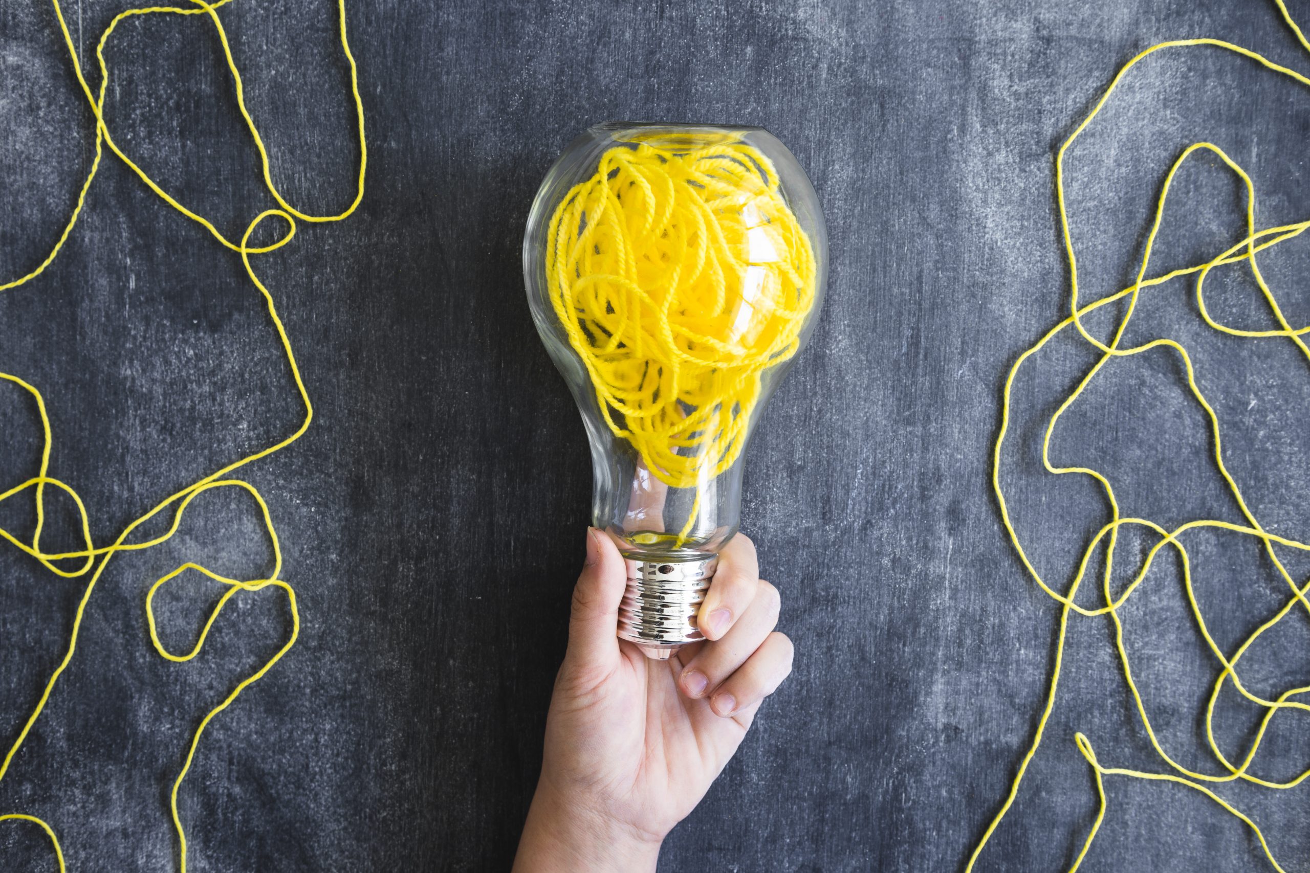 close up hand holding transparent light bulb with yellow yarn blackboard scaled